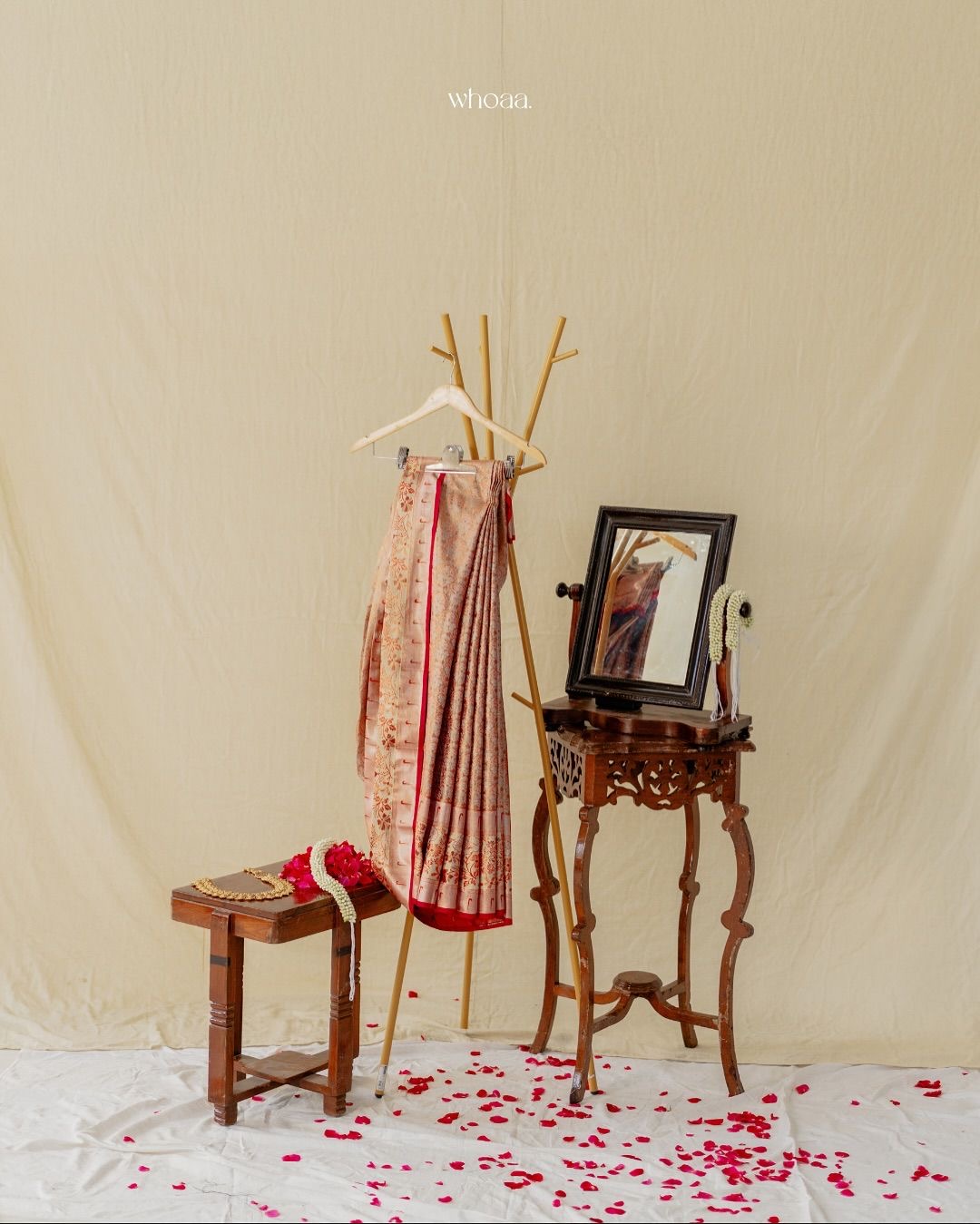 Pink and gold embroidered saree on a wooden hanger beside a carved mirror stand and rose petals